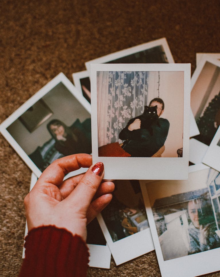 A hand with red nails holds a vintage Polaroid among scattered photos on carpet.