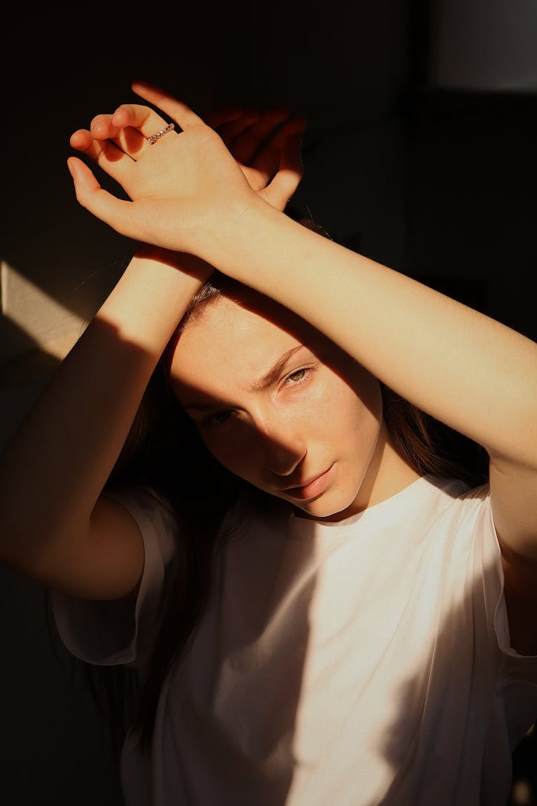 A young woman in a white shirt, hands raised, with light casting shadows indoors.