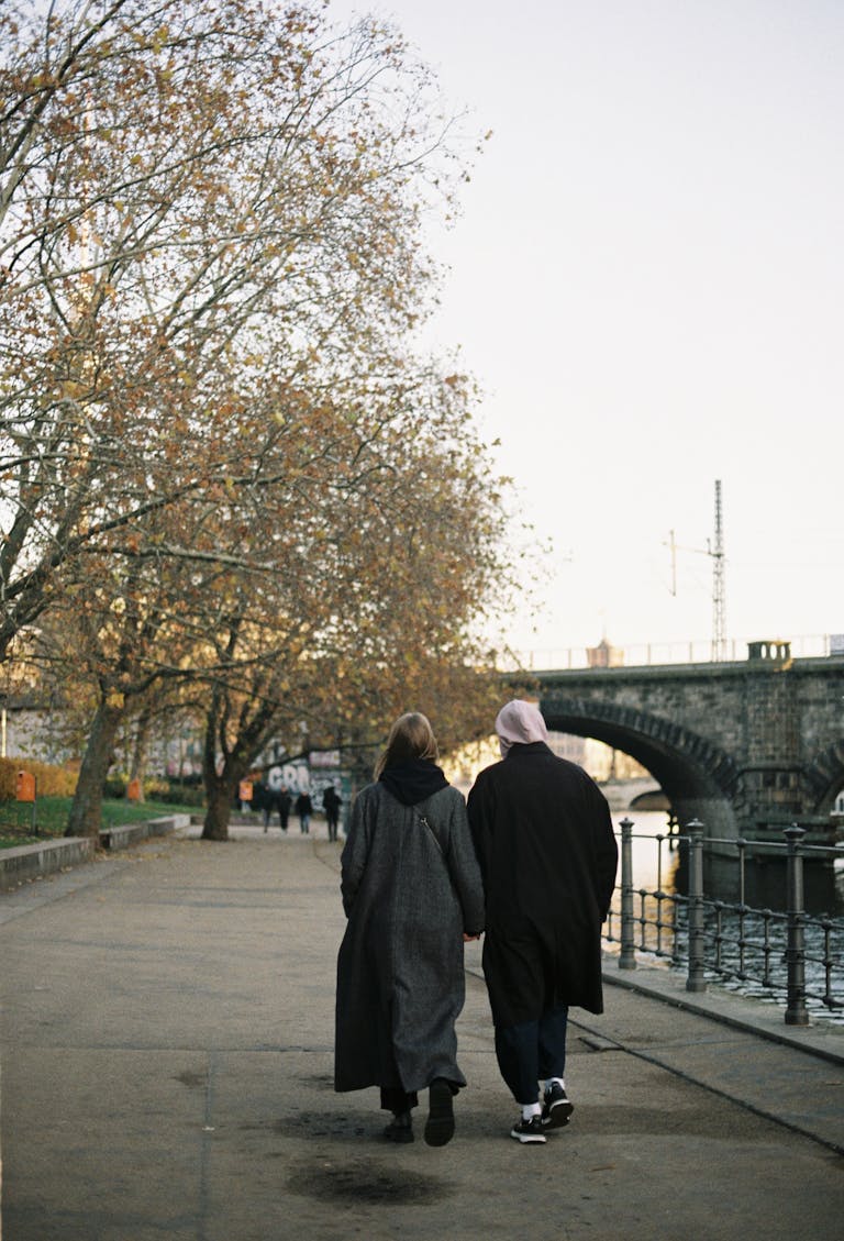 Couple walking hand in hand along a tree-lined riverside path in autumn.