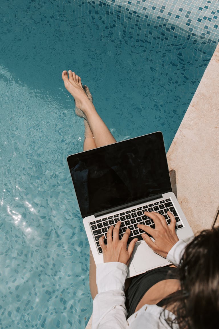 Woman working remotely on a laptop by the poolside, enjoying summer outdoors.