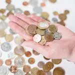A hand holding a variety of coins with more scattered on a white surface, symbolizing currency and finance.