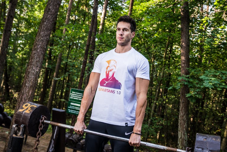 A man performs a weightlifting exercise in a forest park, showcasing fitness and strength outdoors.