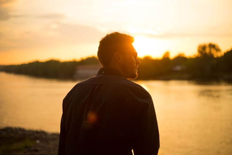 A man stands by the river as the sun sets, creating a serene silhouette.