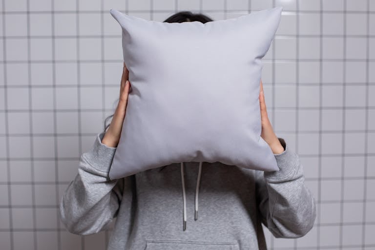 A person in a grey hoodie holds a soft grey pillow against a tiled background.