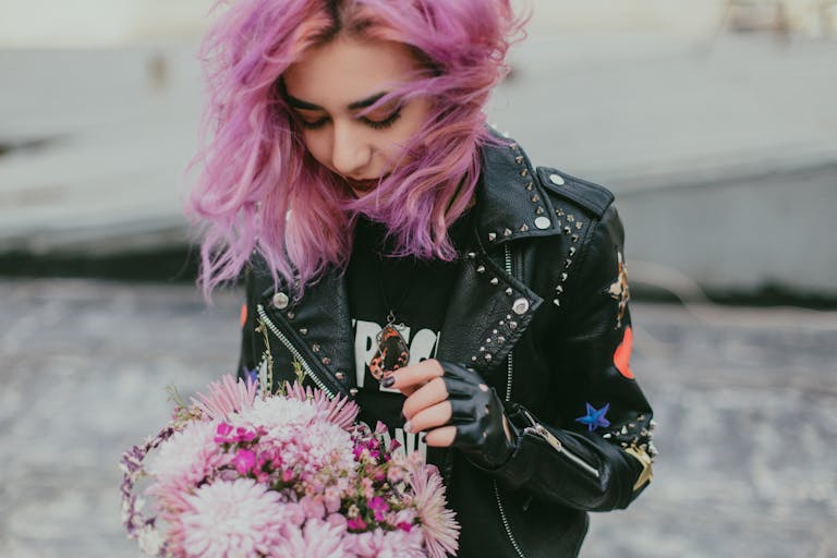 A stylish young woman with purple hair holding a bouquet, wearing a black leather jacket outdoors.