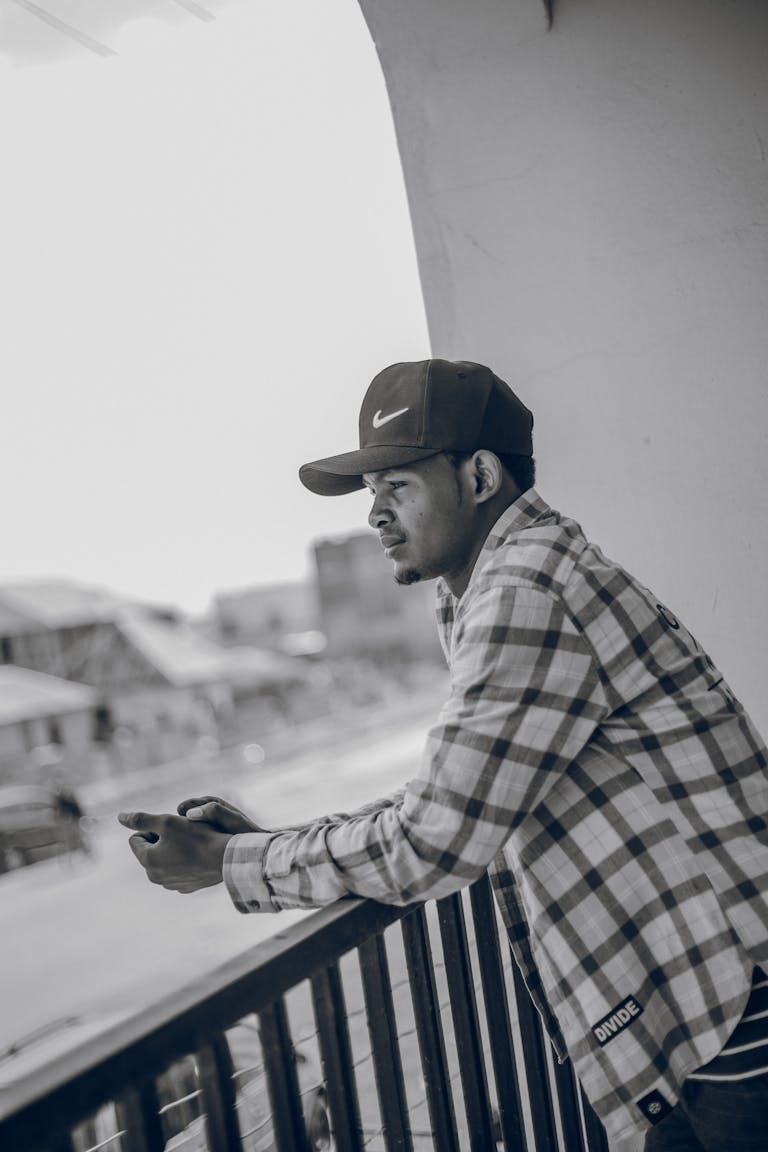 A young man in a plaid shirt leans on a balcony railing, gazing outward.