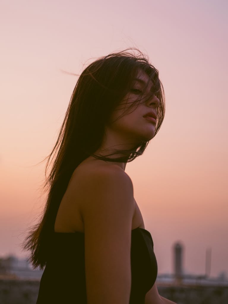 A young woman stands at sunset, hair blowing gently in the wind, evoking a serene and artistic mood.