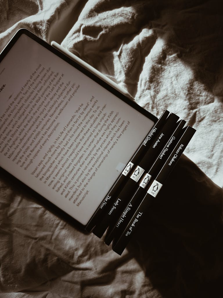 An ebook reader and hardcover books on rumpled bed casting shadows in sunlight.