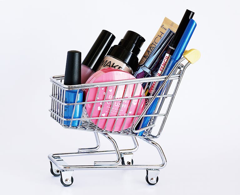 Miniature shopping cart filled with a variety of cosmetics on a white background.