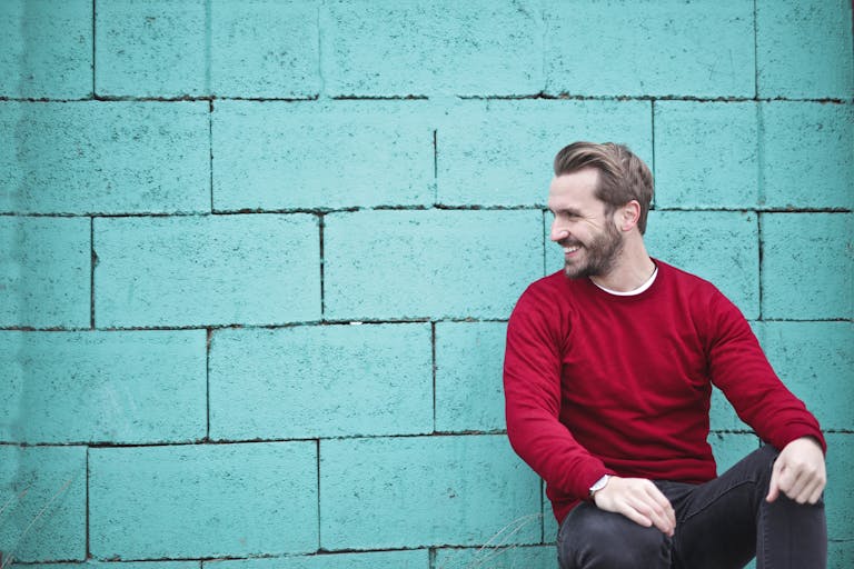 A cheerful man in a red sweater sits against a turquoise wall, enjoying a moment of happiness and laughter.