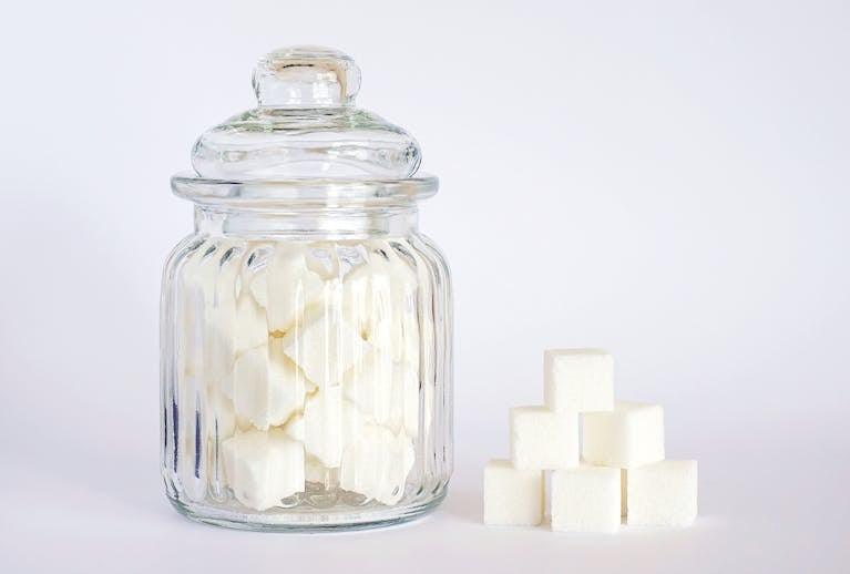 A clear glass jar filled with sugar cubes next to stacked cubes on a white background.