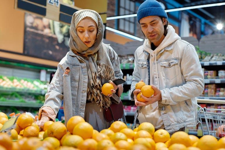 A couple wearing casual attire selects oranges in a busy supermarket aisle.